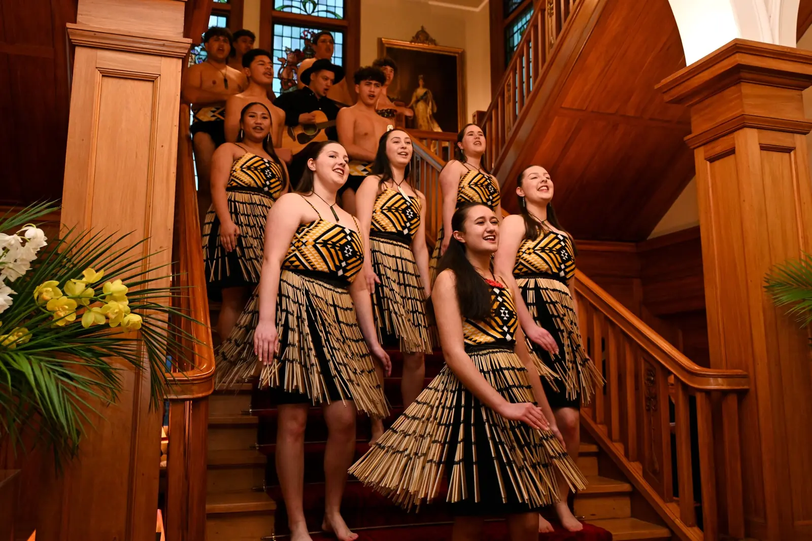 A kapa haka group in woven flax skirts singing on a timber staircase, with more performers standing behind them (not at Auckland Museum)