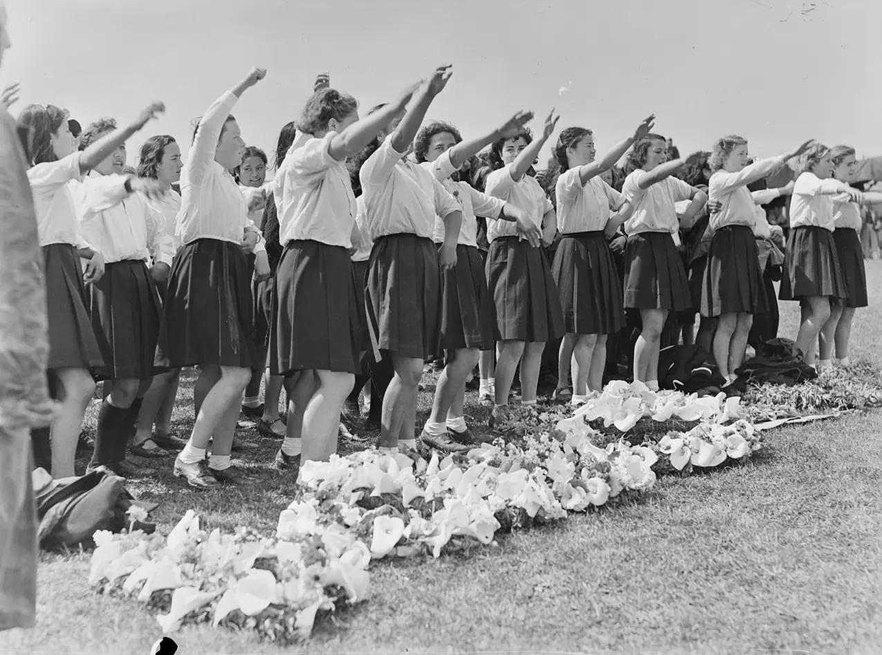 A group of schoolgirls in white blouses and dark skirts standing in lines on grass with one arm raised, bouquets of lilies on the ground before them