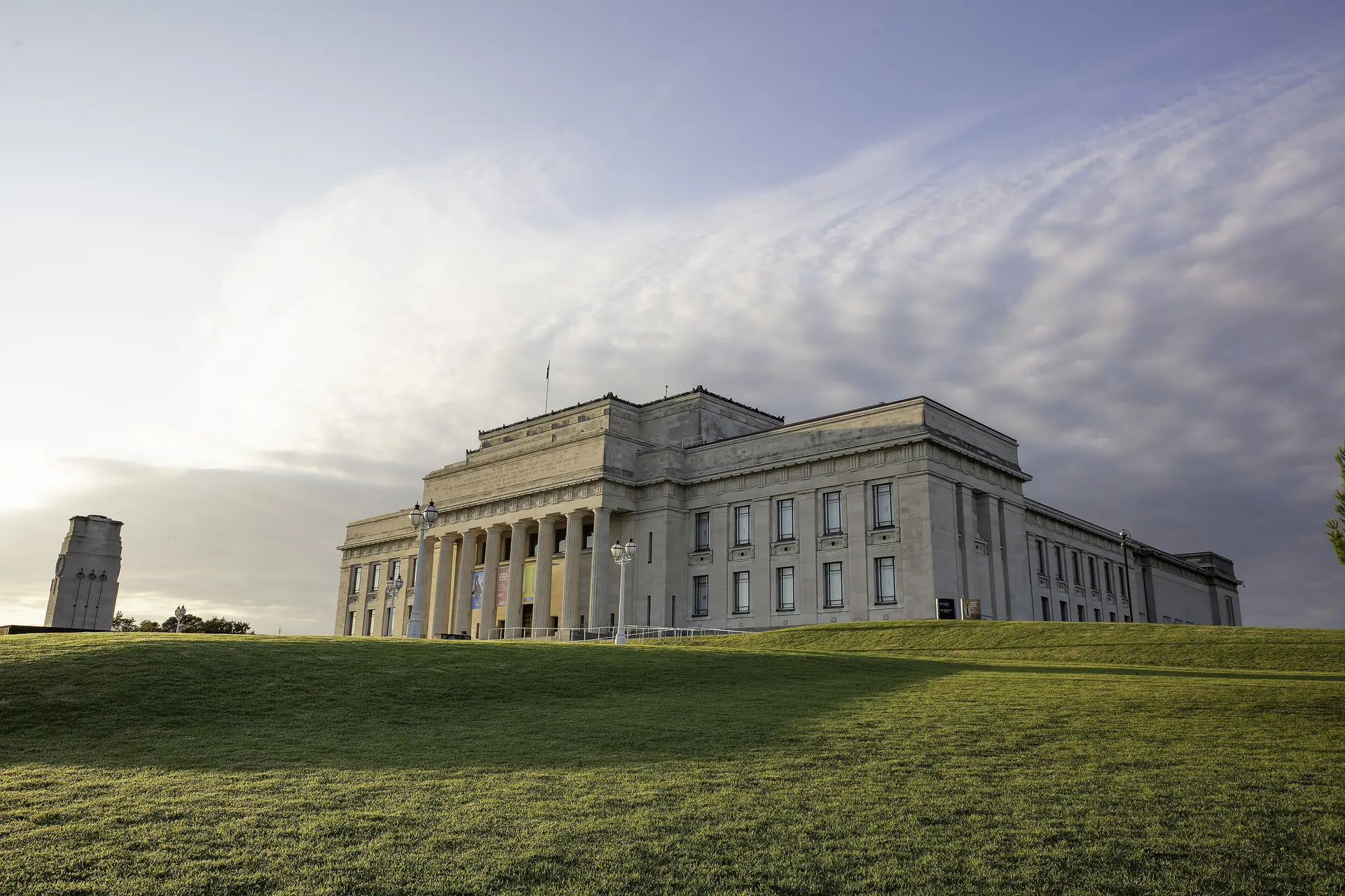 Auckland War Memorial Museum at dusk, its columned neoclassical front lit by low sun above a grassy slope in the Auckland Domain